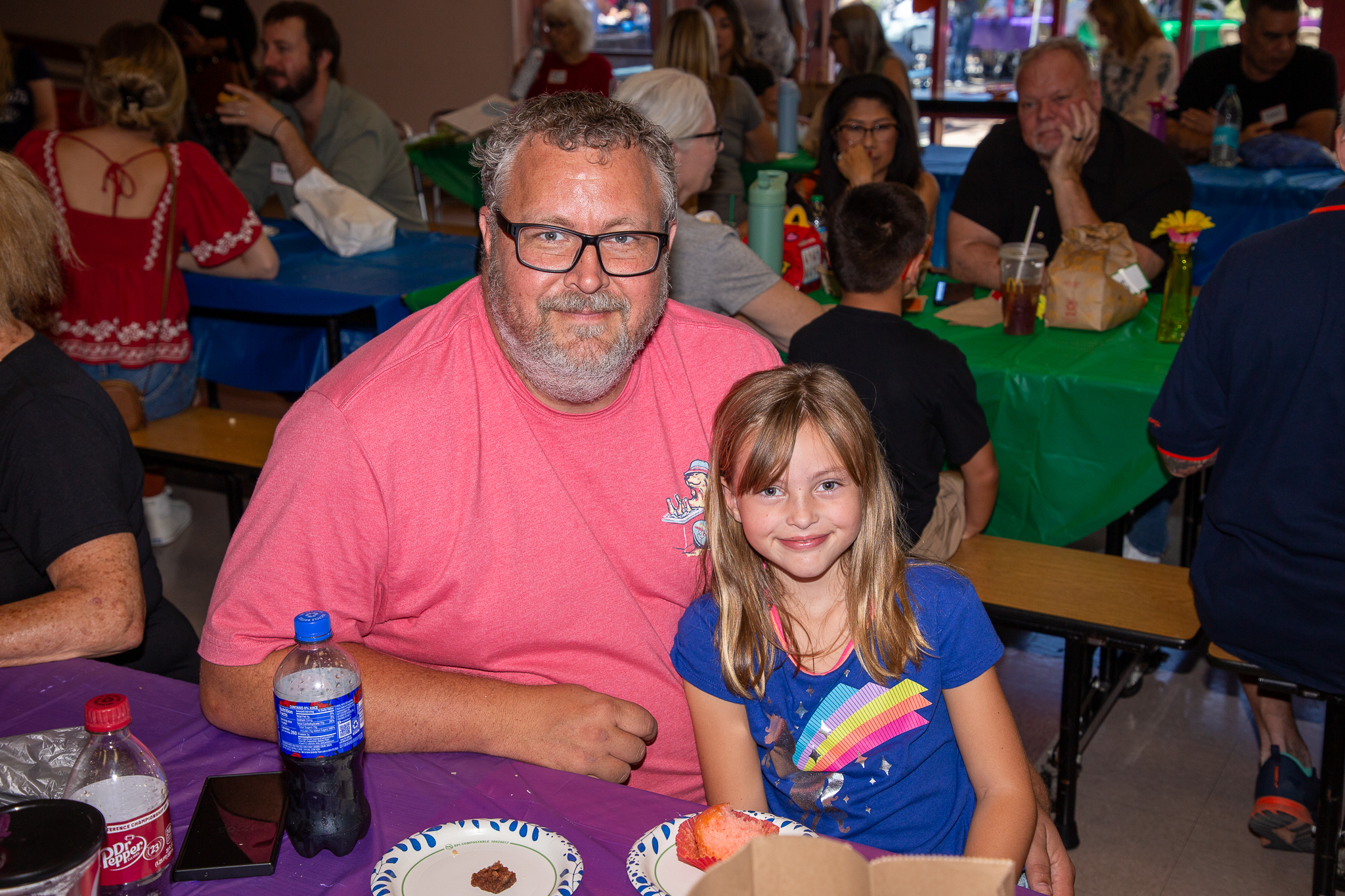 A grandpa in a pink shirt and glasses smiles with his granddaughter in a blue shirt