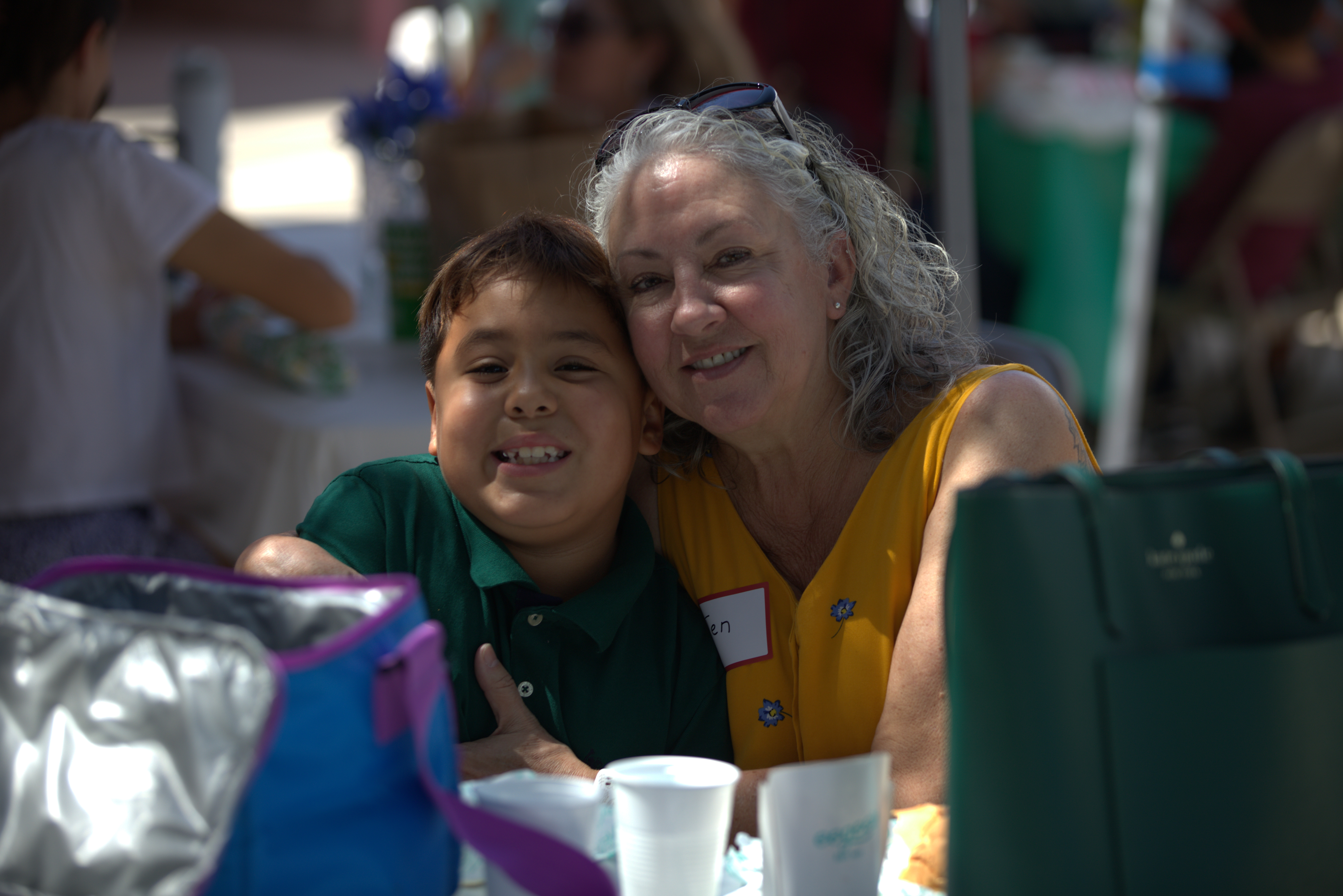 A grandma in a yellow shirt smiles with her grandson in a green shirt