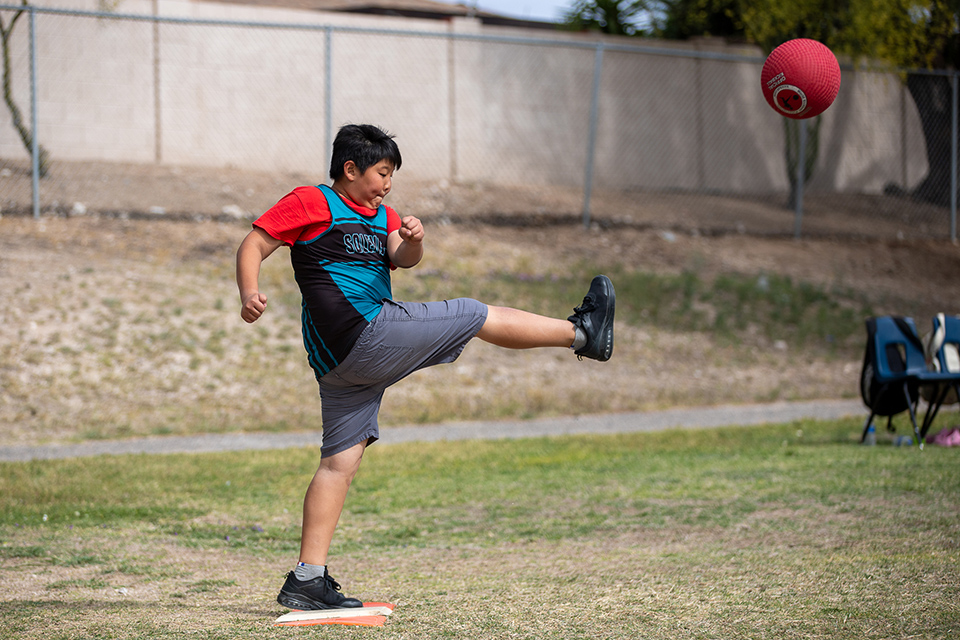 A boy in a teal jersey and red shirt kicks the red ball hard